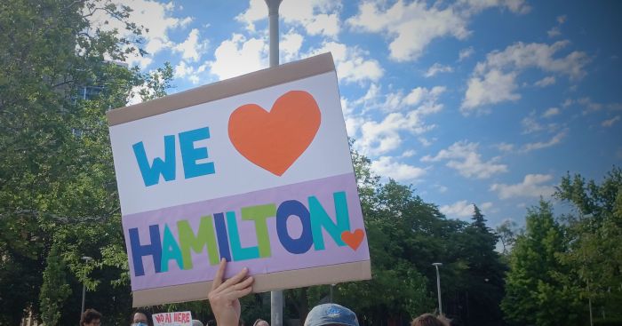 protest sign reading 'We (heart) Hamilton' rendered in bright construction paper, held up outside City Hall on June 16th, 2026